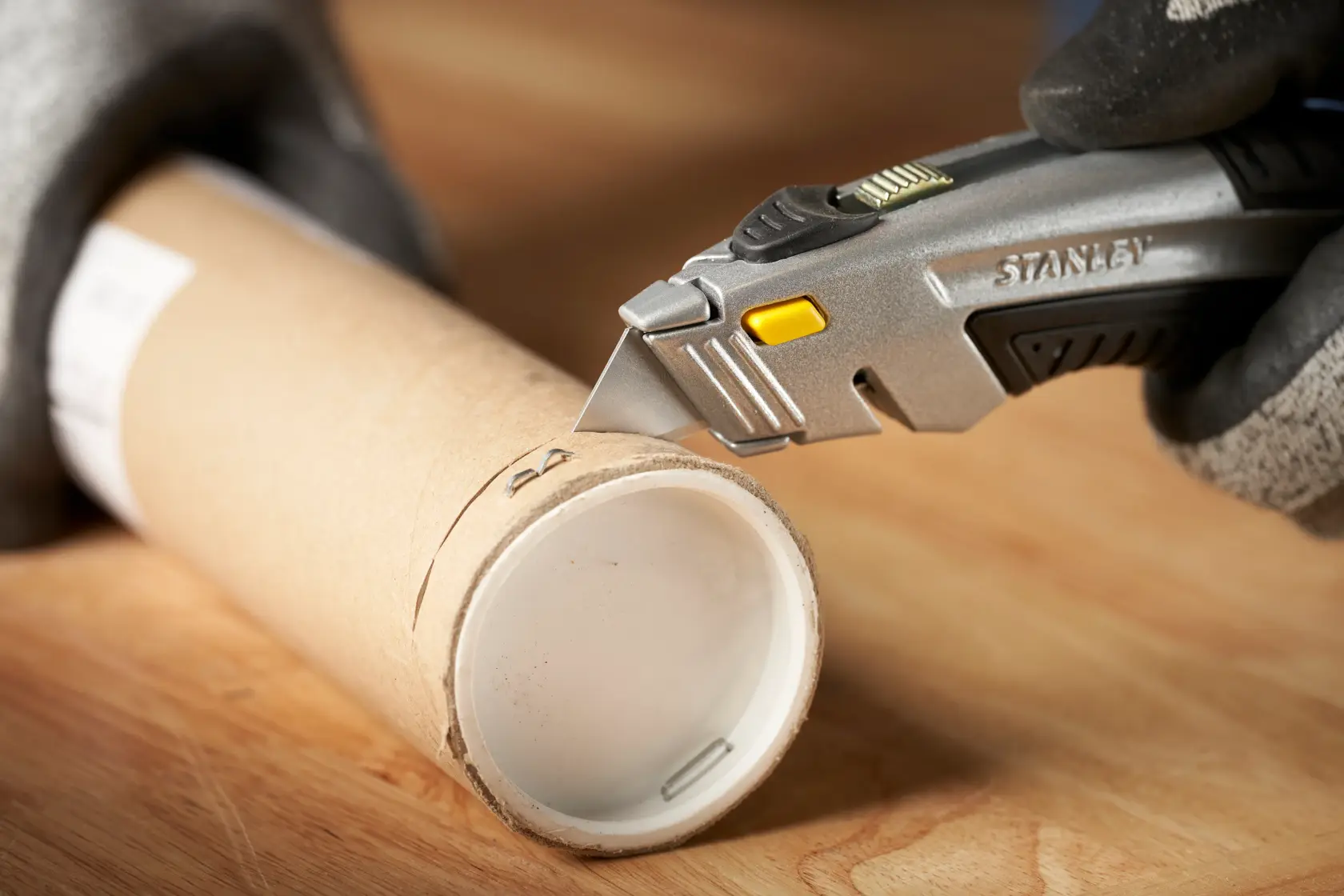 Photo of STANLEY 0-10-788 utility knife cutting cardboard tube on wood surface, close-up side angle.