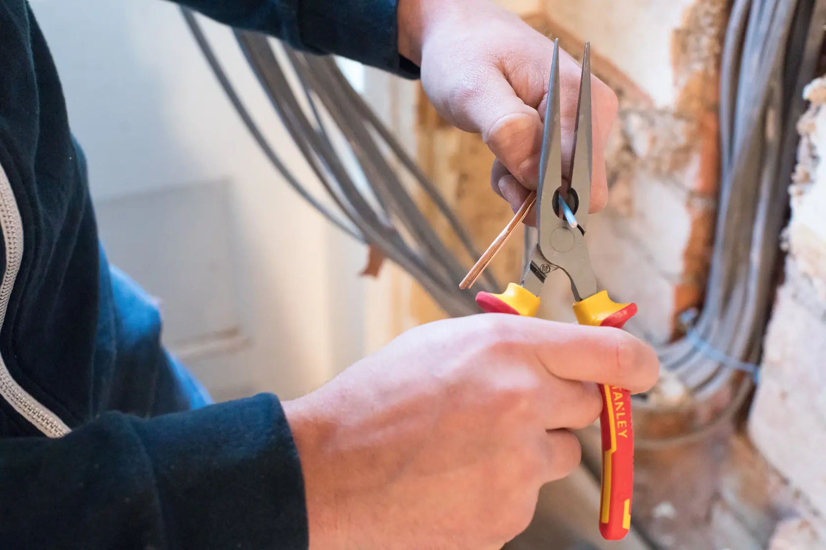 STANLEY 0-84-007 long nose pliers held in hand, gripping wire near wall, tool with yellow and red handles.