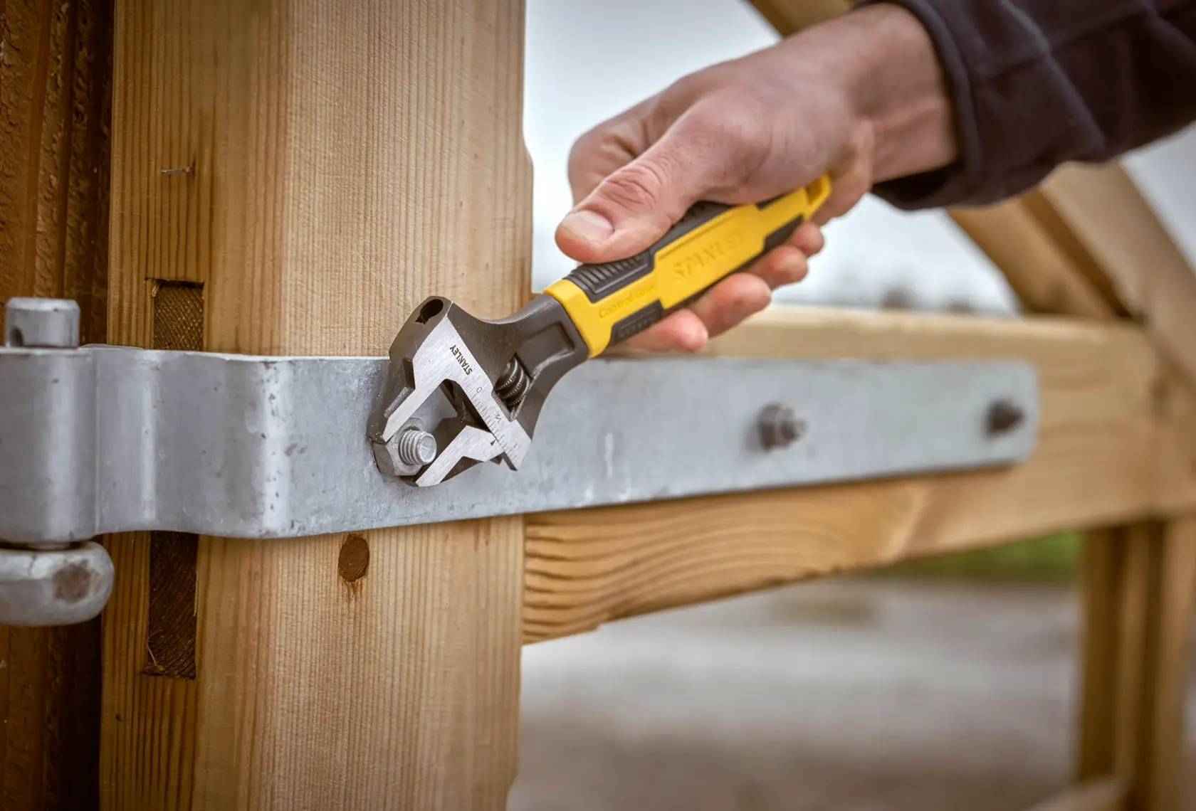 Stanley 0-90-949 adjustable wrench tightening a bolt on a metal hinge attached to a wooden gate, close-up.