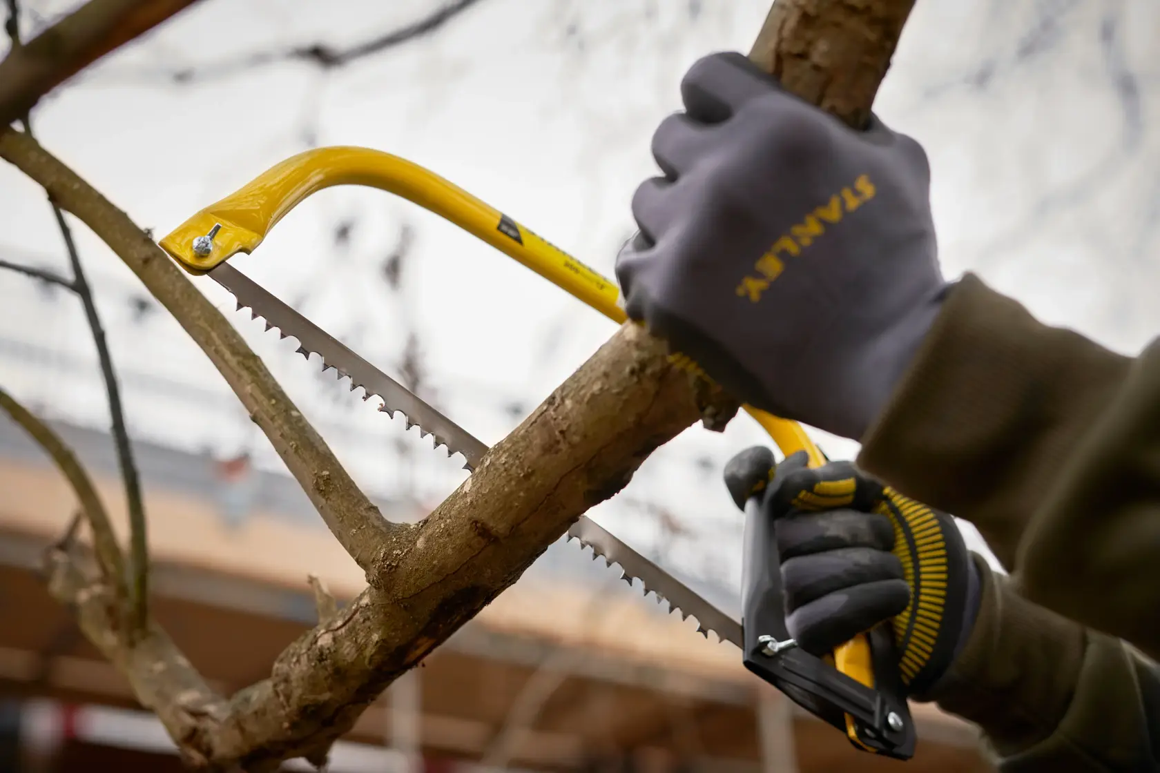 STANLEY 1-20-447 hand saw cutting tree branch with gloved hands, yellow frame, close-up outdoor photo.
