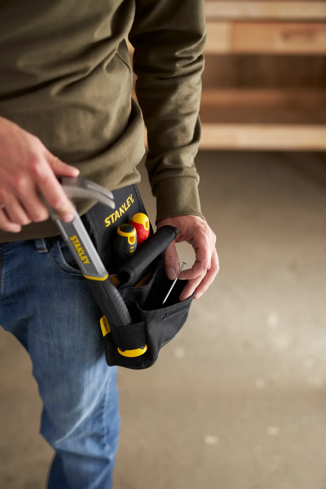 Photo of a person wearing a STANLEY tool belt pouch SKU 1-93-329 holding hand tools, indoor workshop setting.