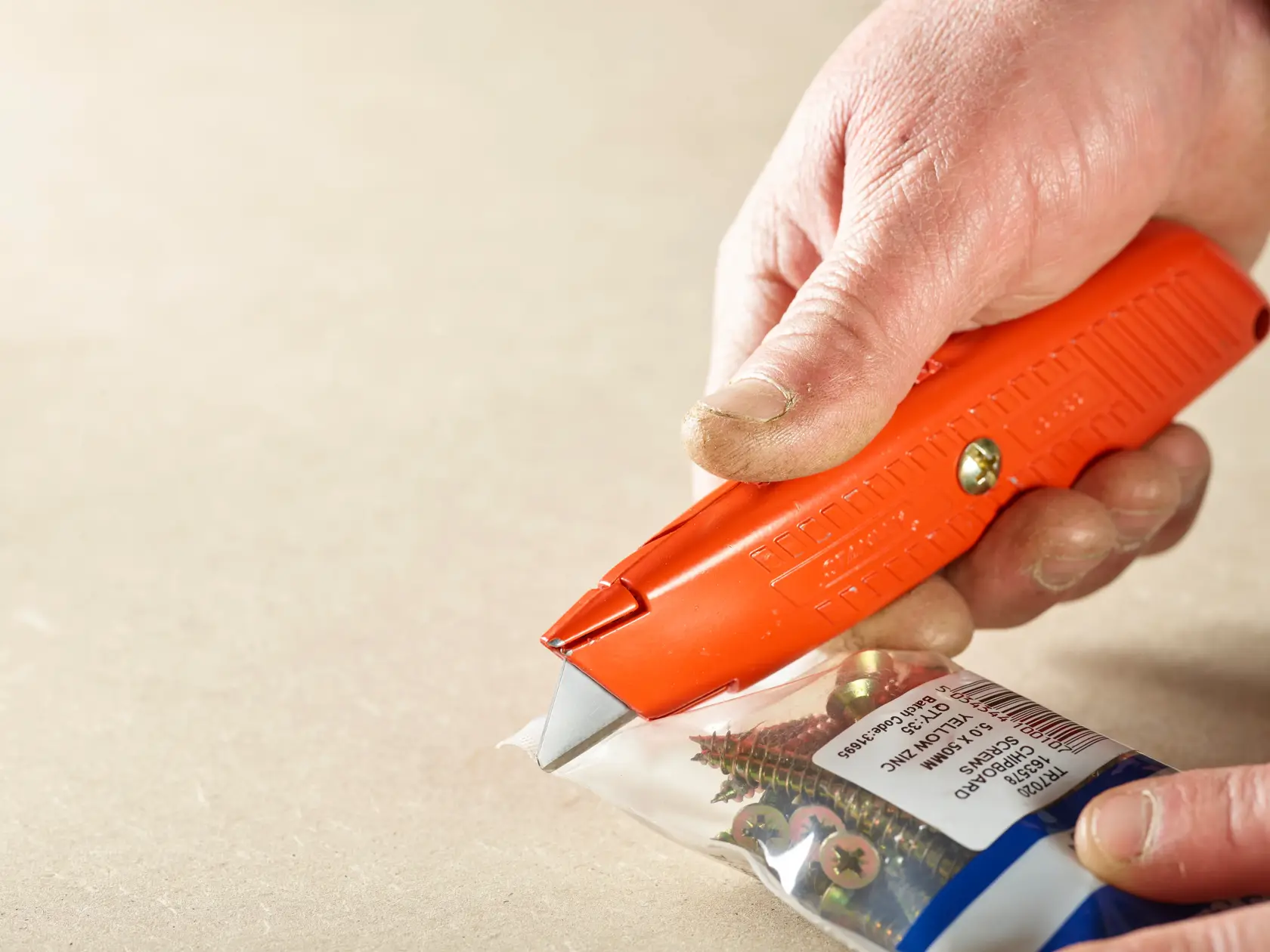 STANLEY 2-11-987 utility knife cutting plastic screw packaging, close-up showing orange handle and blade.