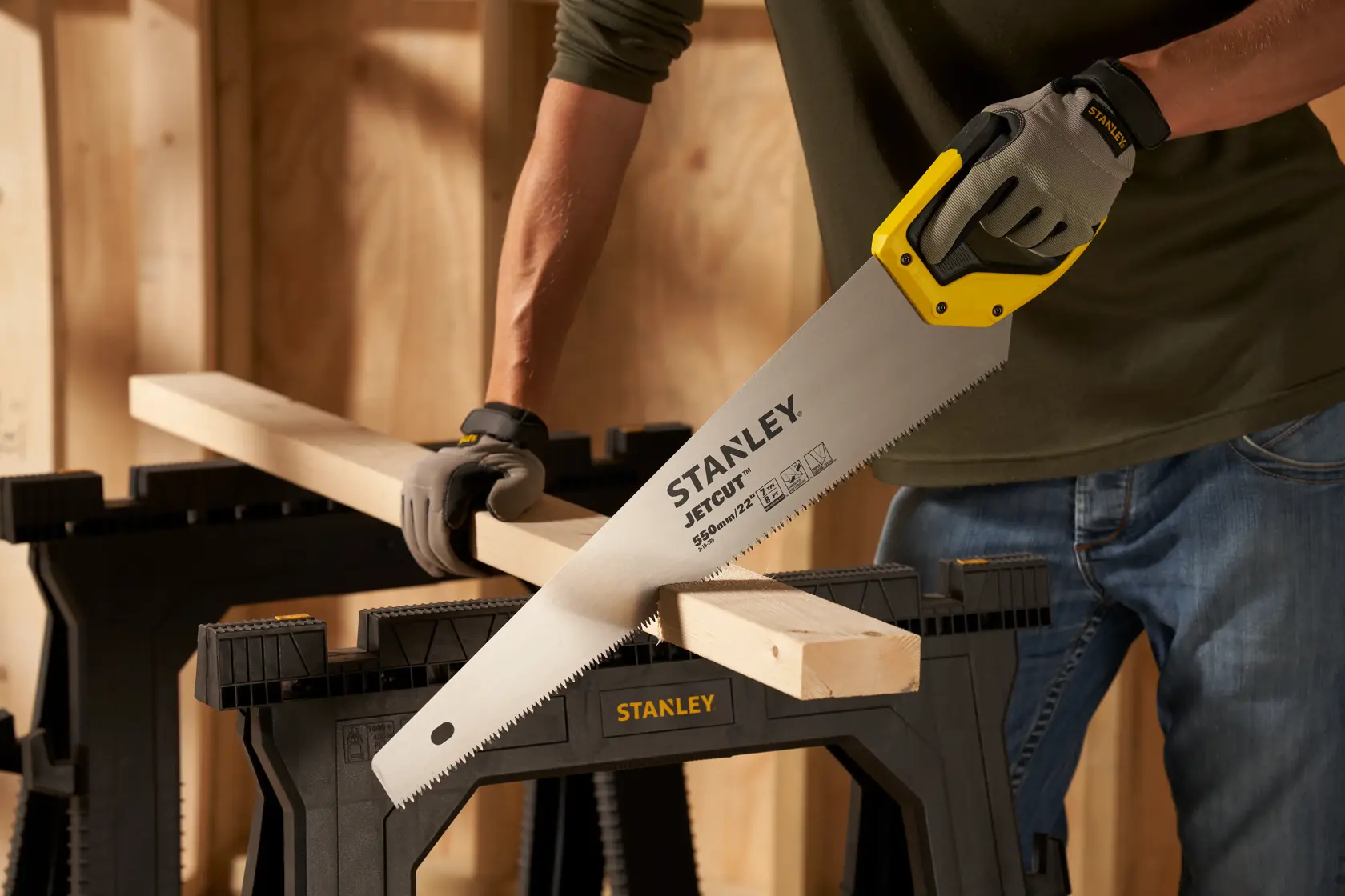 Stanley 2-15-289 hand saw with yellow handle cutting wood on black sawhorse, workshop background.