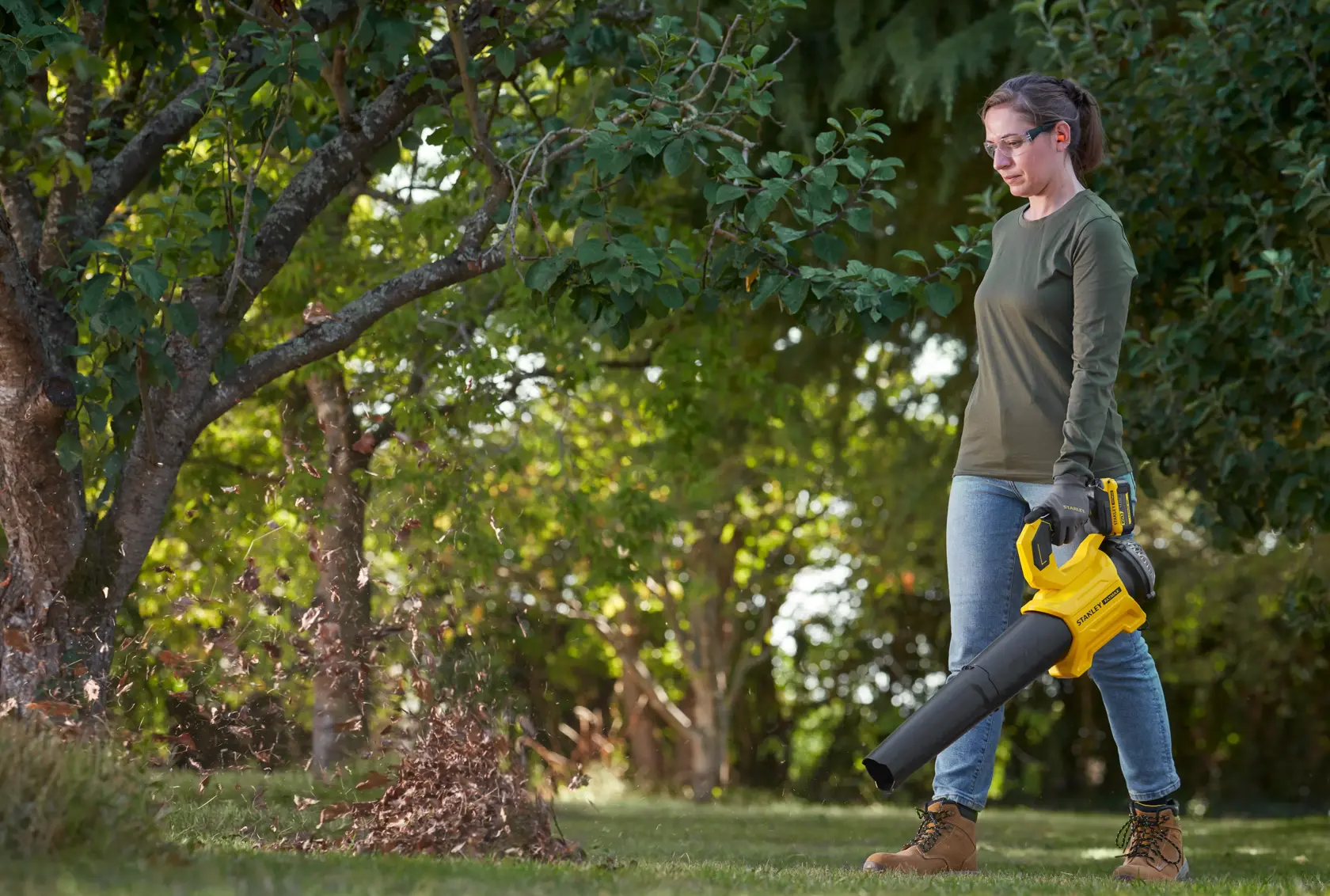 STANLEY SFMCBL730B leaf blower in use outdoors, yellow and black design, blowing leaves under a tree.