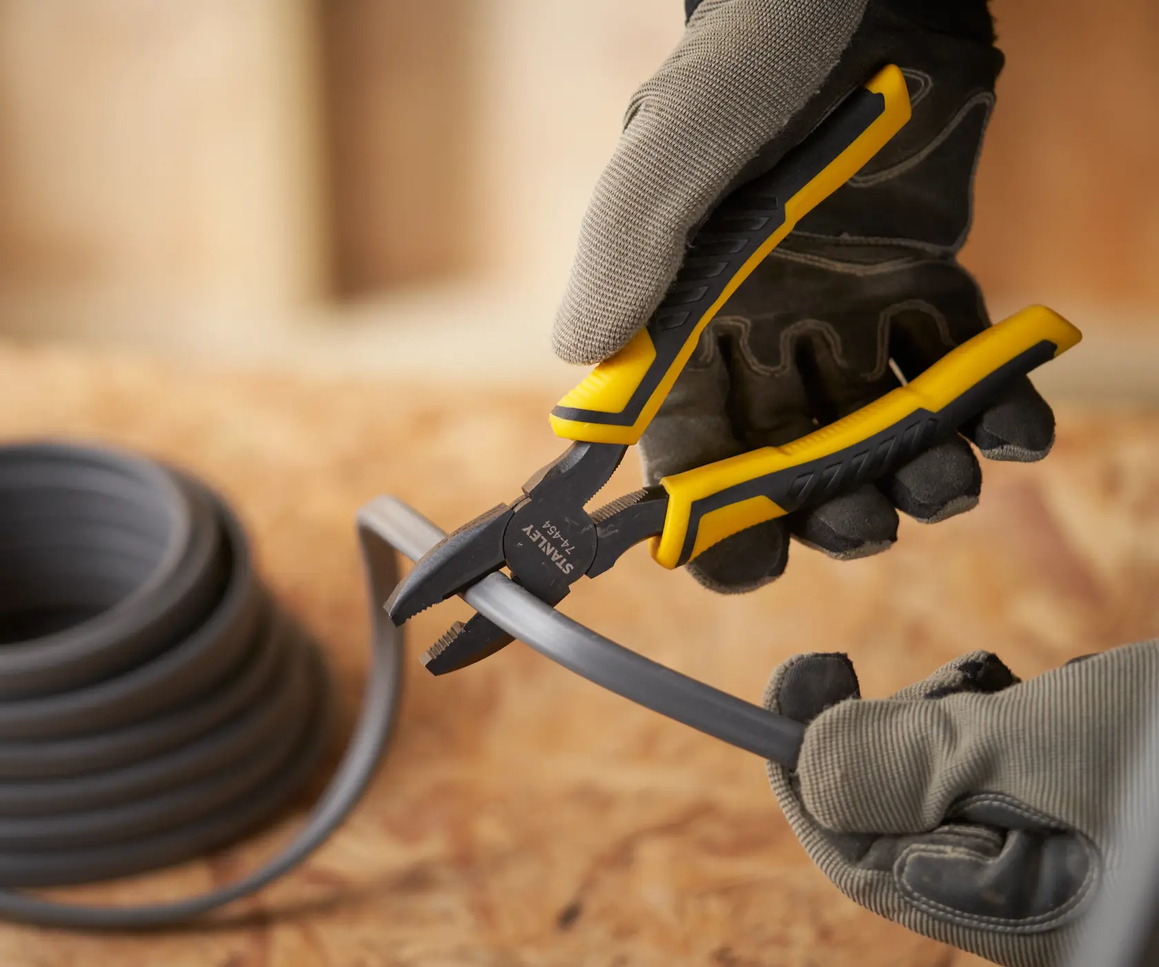 STANLEY STHT0-74456 pliers with yellow and black handle cutting black cable, worn by gloved hand, close-up.