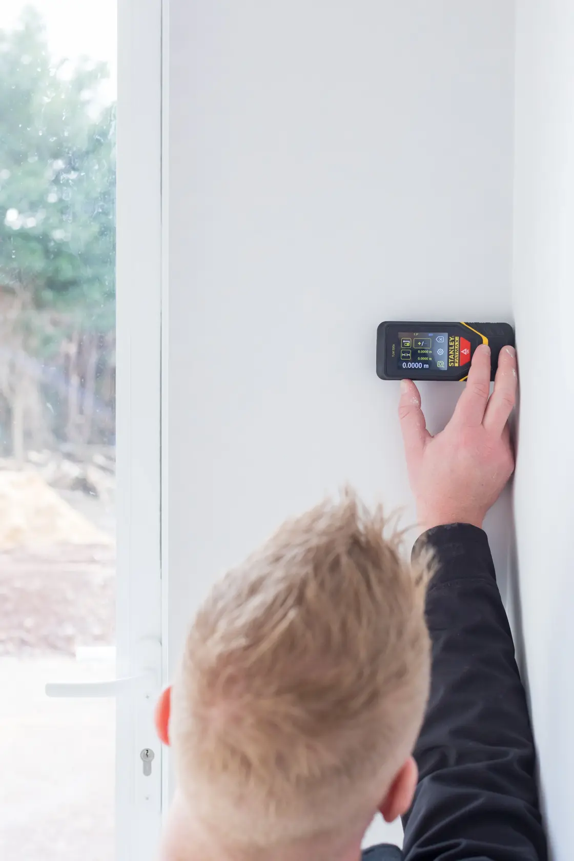 STANLEY STHT1-77140 laser distance measurer held against white wall by person, side angle.