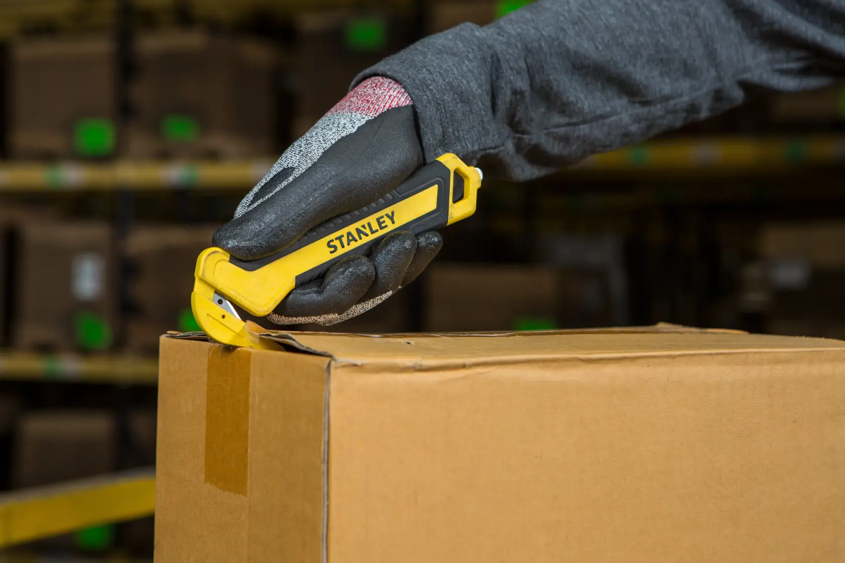 STANLEY STHT10356-0 safety utility knife cutting a cardboard box, handheld, warehouse background.