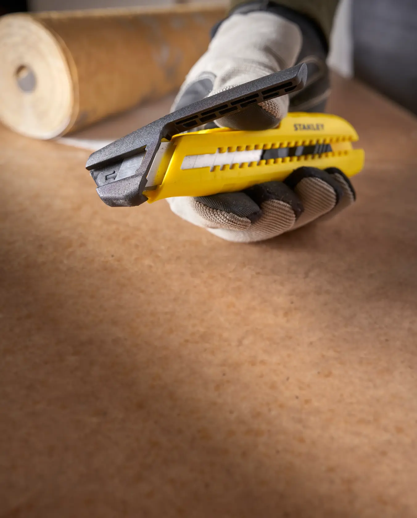 Photo of STANLEY STHT10489 yellow utility knife held in gloved hand on a work surface, roll of material in background.