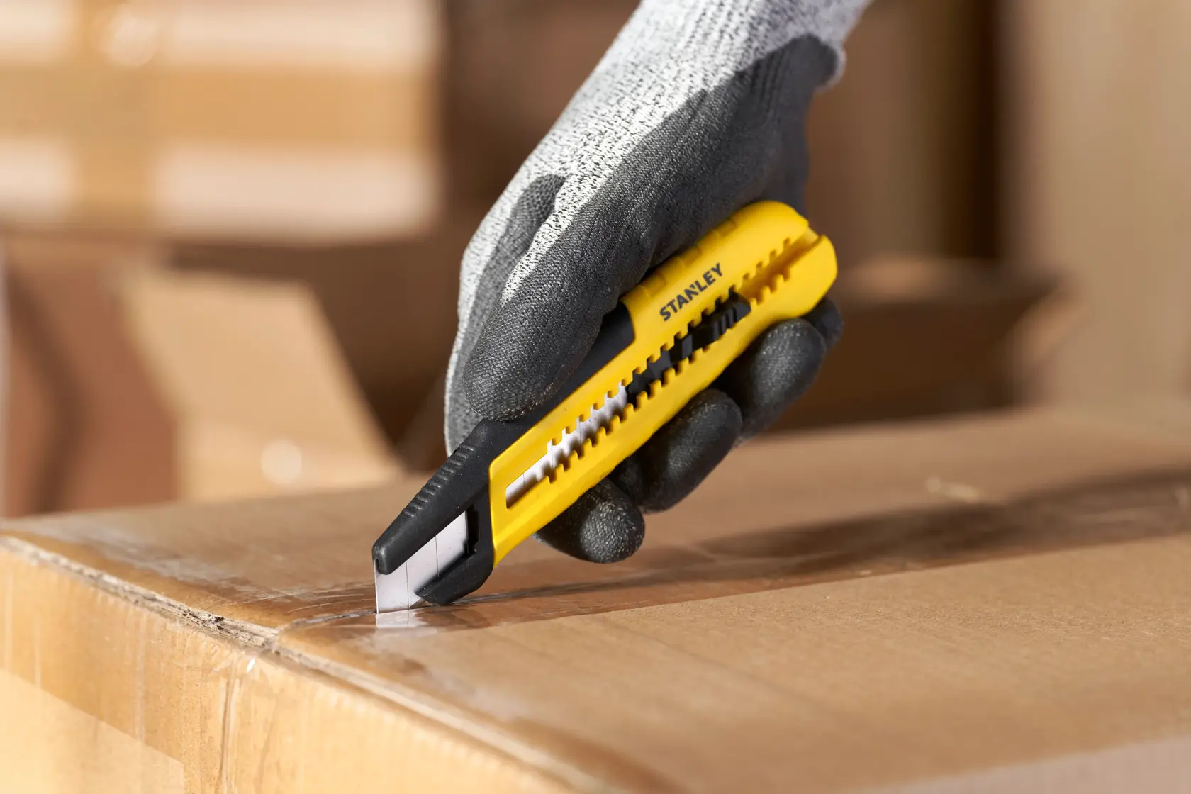 Photography of STANLEY STHT10489 utility knife cutting a cardboard box, held by gloved hand.