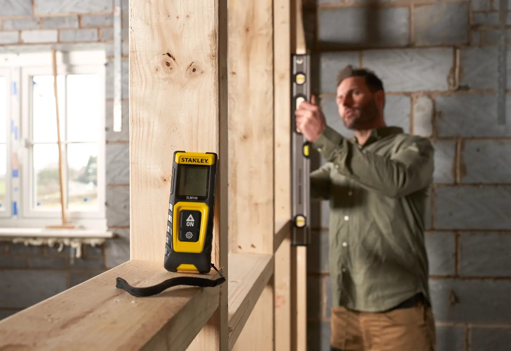 Photo of STANLEY STHT77100-0 laser distance measurer on wooden frame at construction site, with person using level in background.