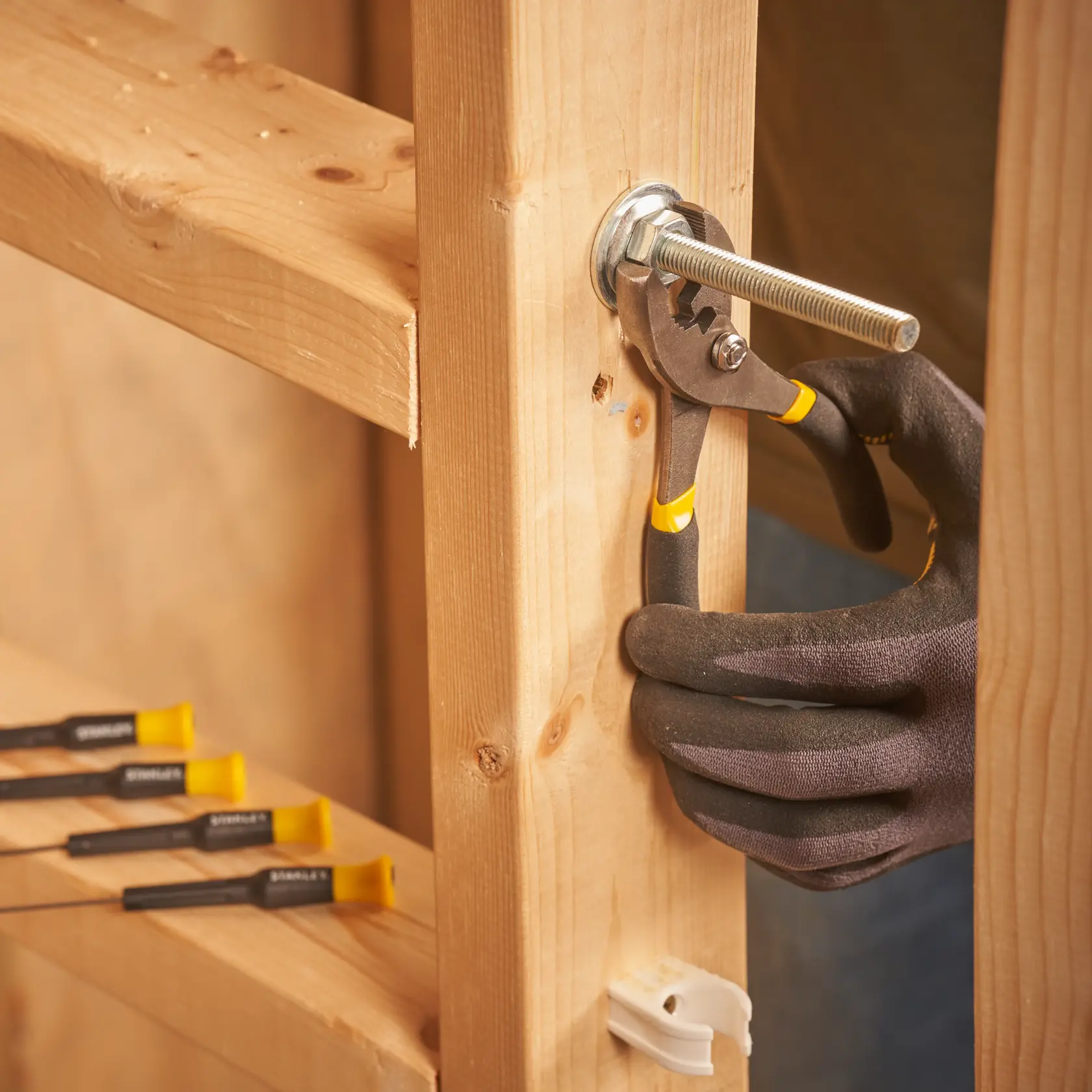 STANLEY STMT0-74101 pliers gripping a bolt on wooden frame, close-up photo, hand in black glove.