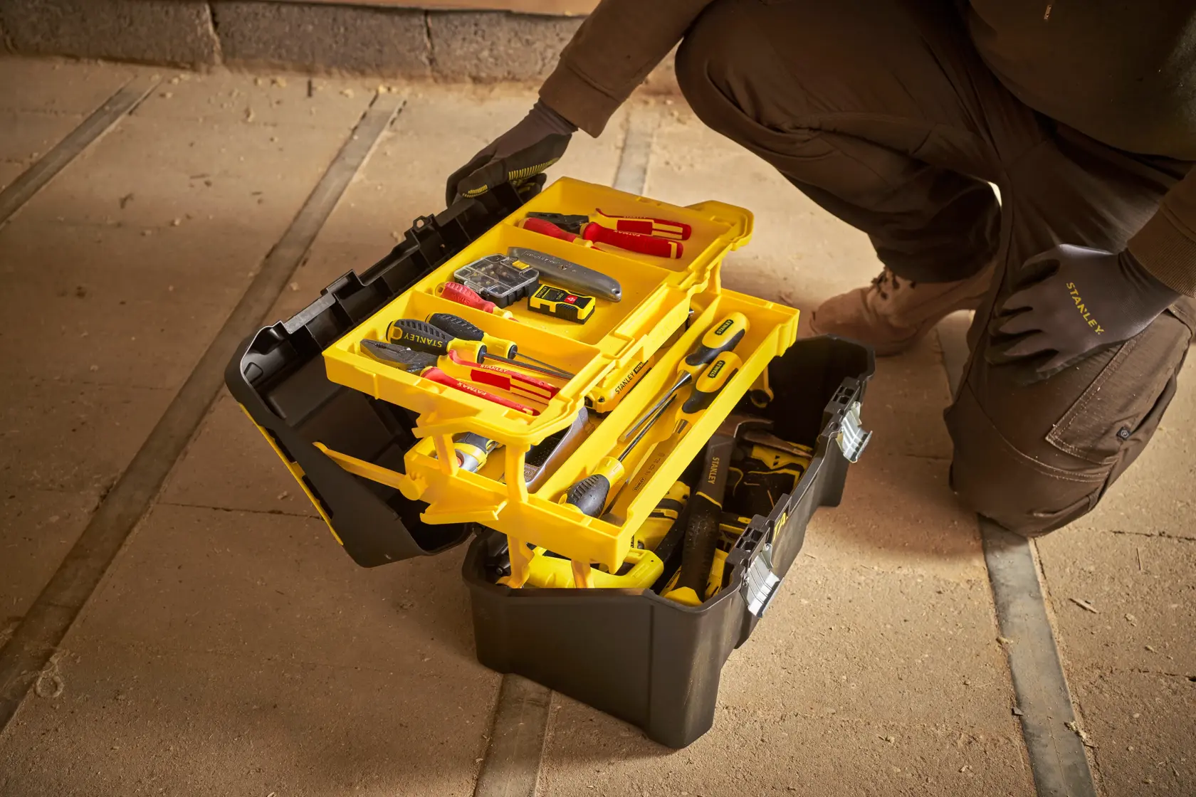 STANLEY STST83397-1 black and yellow tool box with open trays showing assorted hand tools on workshop floor.