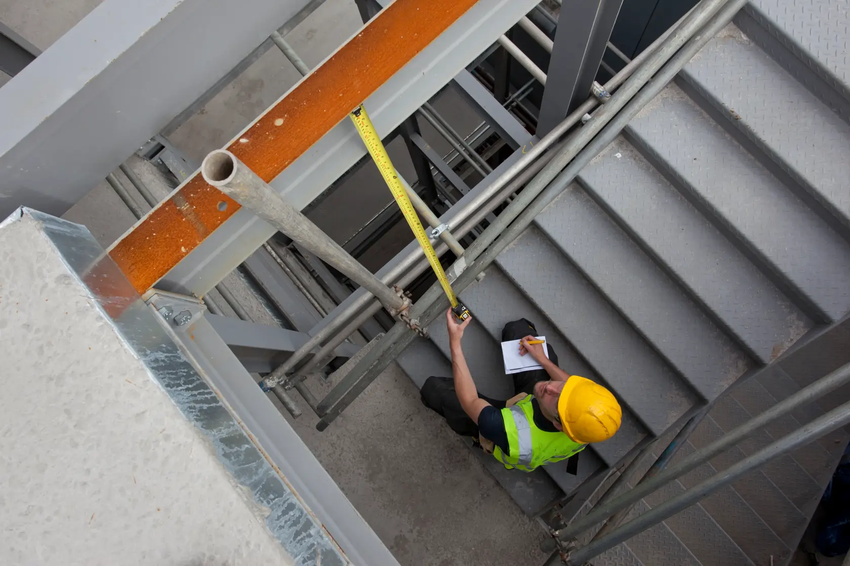 STANLEY XTHT0-33671 tape measure used by worker in safety gear measuring metal stairway, overhead angle.